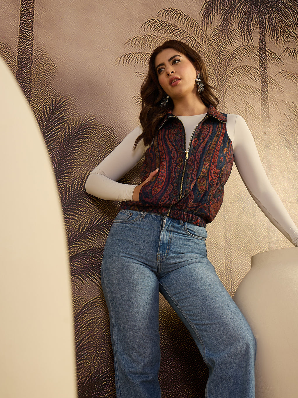 Woman wearing a patterned vest and blue jeans against a decorative wall.