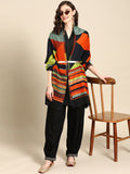 A woman standing next to a wooden chair, wearing a multicolored geometric woven design stole with fringed border, paired with black trousers and sunglasses.