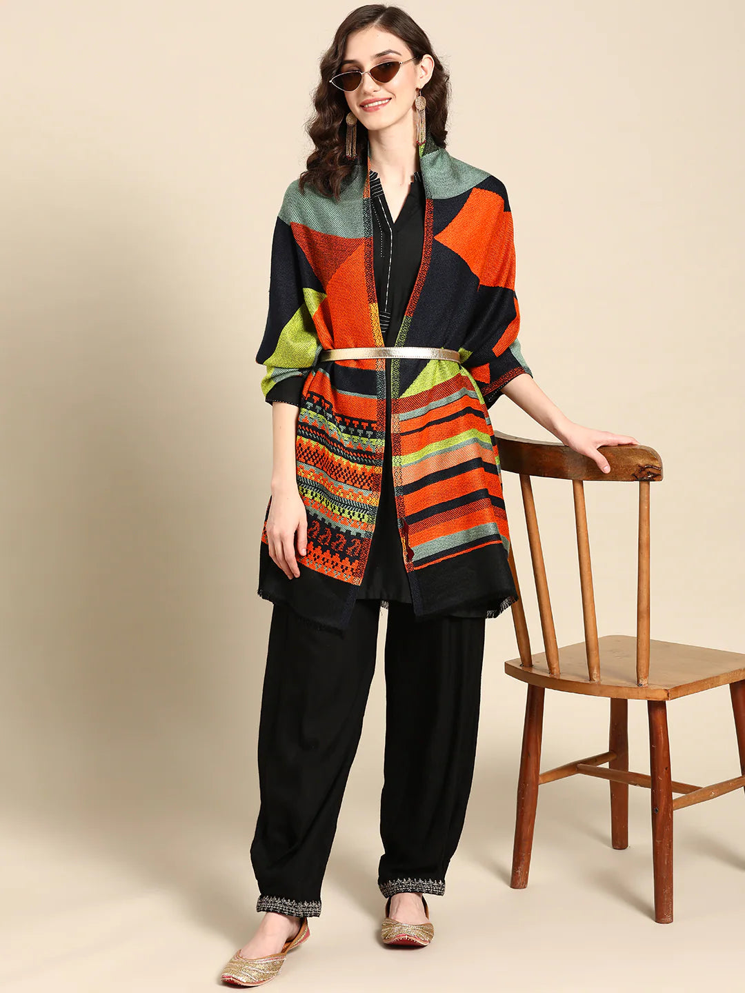 A woman standing next to a wooden chair, wearing a multicolored geometric woven design stole with fringed border, paired with black trousers and sunglasses.